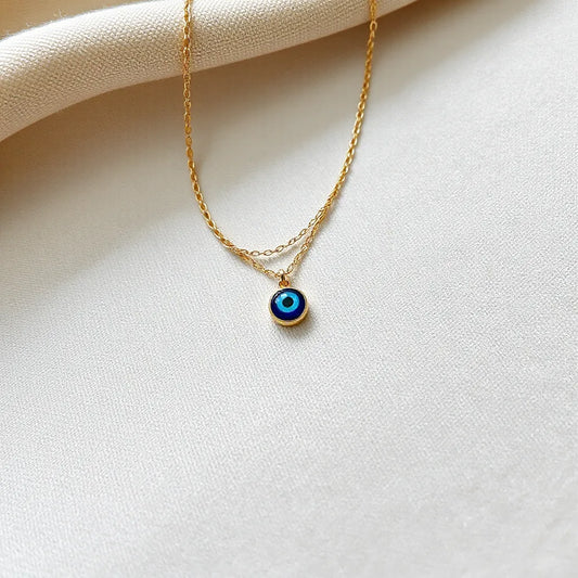 Layered necklaces with small blue evil eye pendants resting on neutral fabric