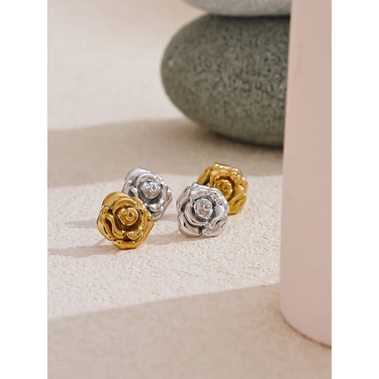 Three rose-shaped earrings in gold, silver, and bronze on a textured surface.