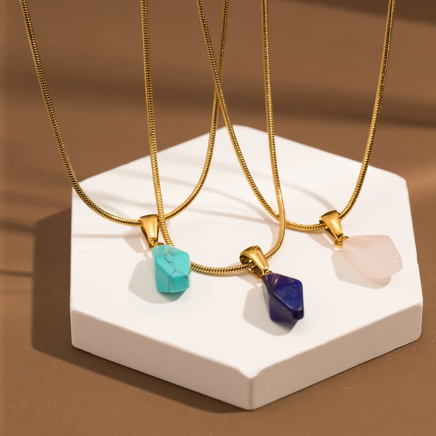 Three gold necklaces with stone pendants on a white stand against a brown background